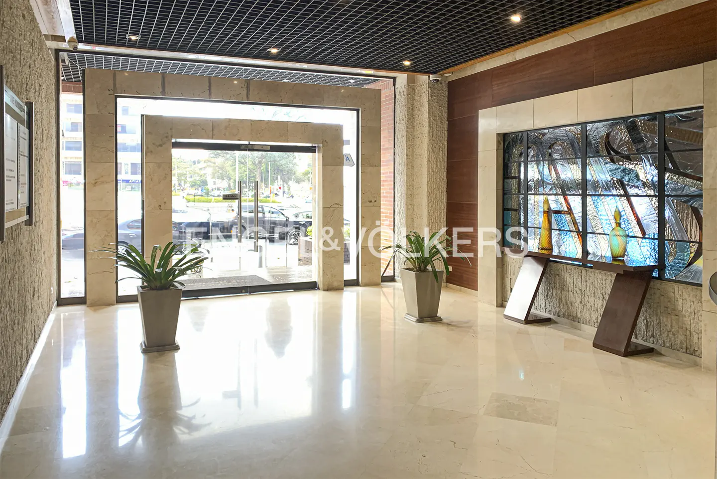 A bright lobby with marble floors, plants, and a stained glass window. An automatic door leads outside.
