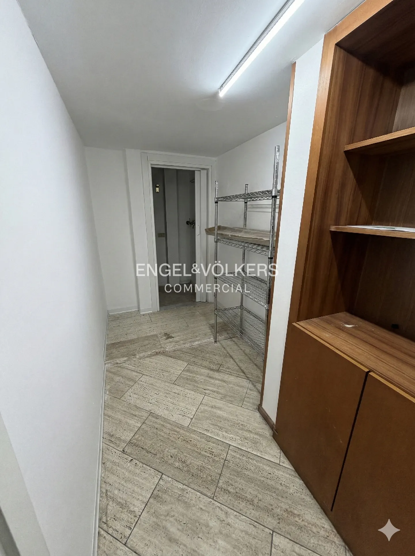 A bright room with beige tile floors, white walls, and a wooden cabinet with shelves. A metal shelving unit stands near an open doorway.