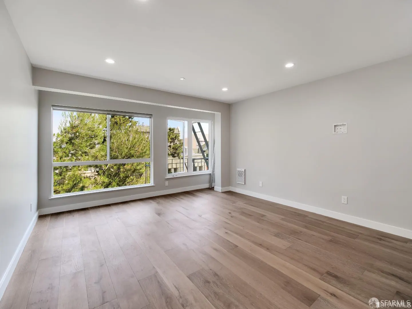 Bright, empty room with hardwood floors, gray walls, and two large windows showing trees and a fire escape.