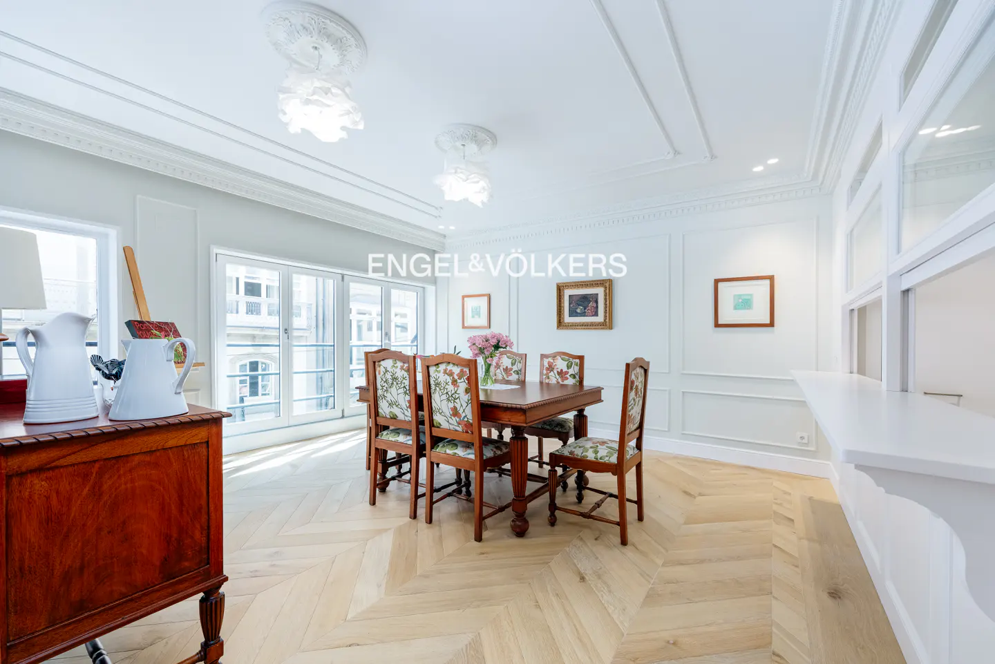 Bright dining room with a wooden table, floral chairs, and herringbone floors. White walls, trim, and ceiling with modern light fixtures.
