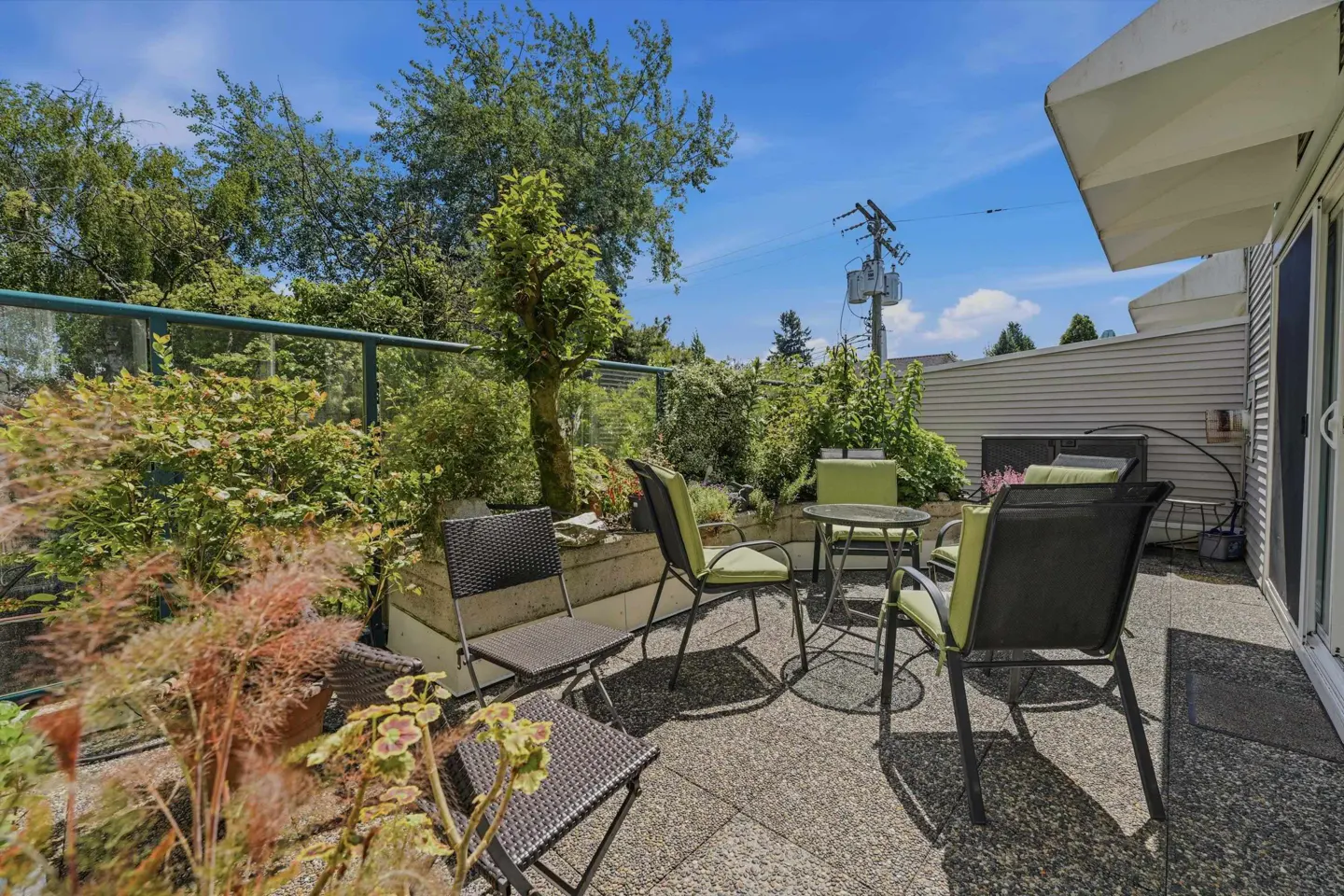 Outdoor patio with table and chairs surrounded by lush greenery, under a bright blue sky.