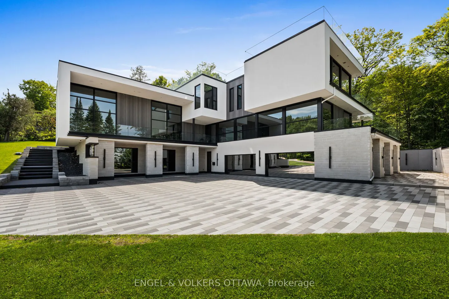 Modern white luxury home with large windows, a paved driveway, and stone stairs, set against a backdrop of green trees.