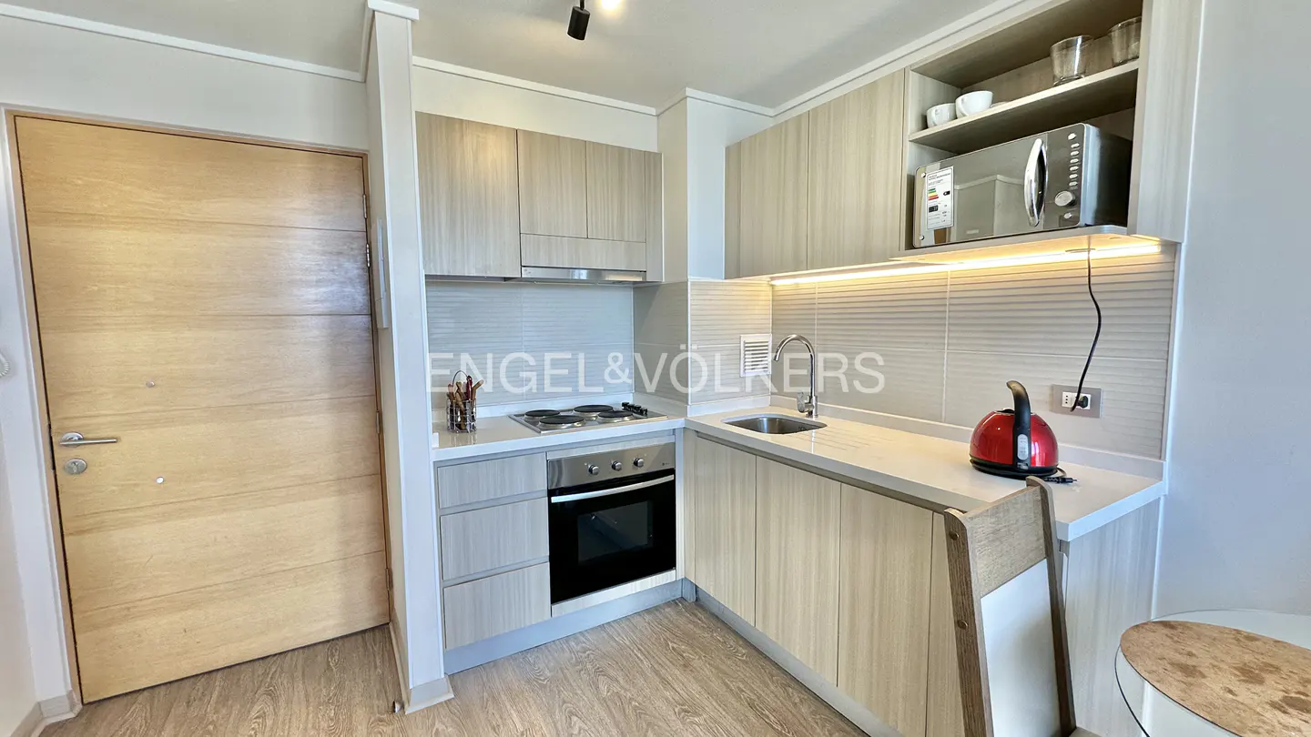 A compact kitchen with light wood cabinets, stainless steel appliances, and a red kettle on the counter. A light wood door is on the left.