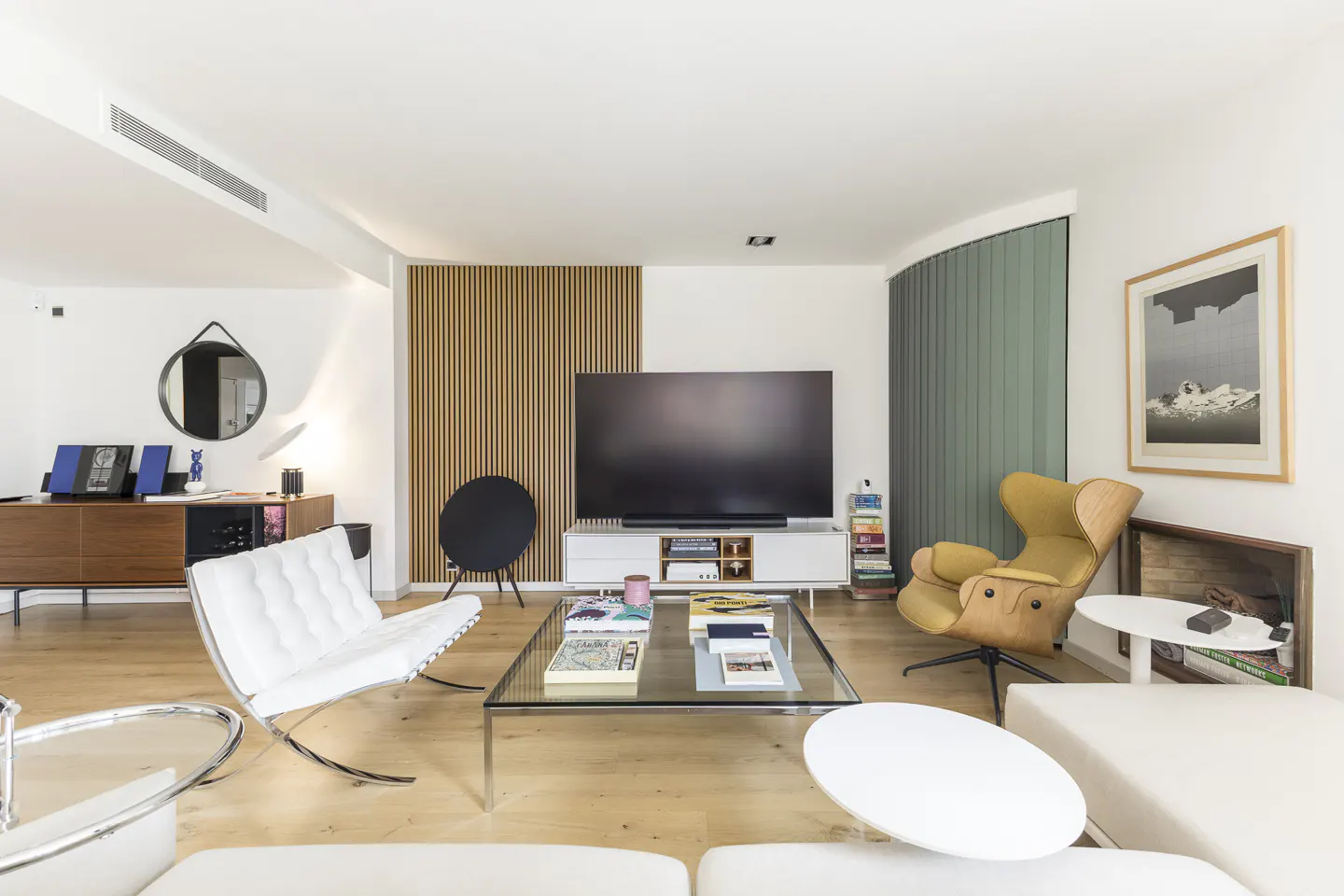 Modern living room with a large TV, white tufted chair, yellow accent chair, glass coffee table, and wood-paneled wall.