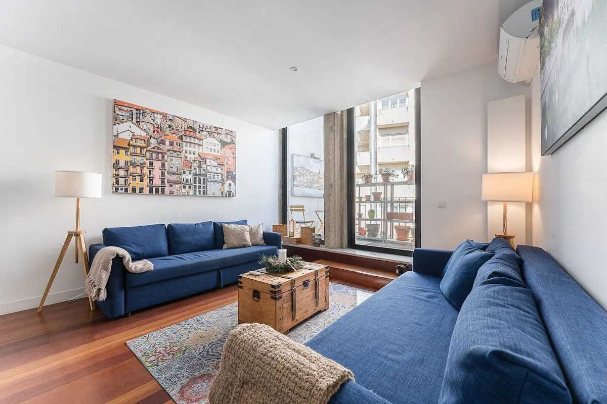 Bright living room with two blue sofas, a wooden chest coffee table, and a colorful city painting on the wall.
