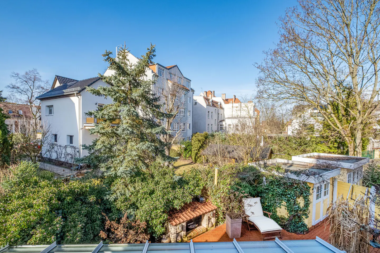 View of a lush garden with a white lounge chair, a yellow shed, and a brick barbecue, surrounded by trees and buildings under a blue sky.