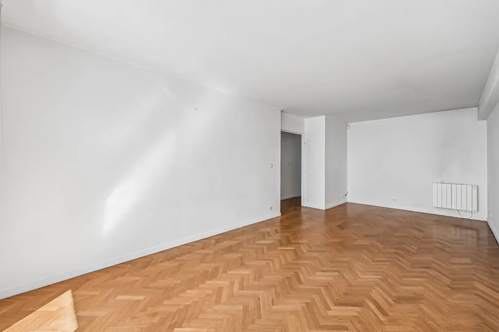 Bright, empty room with white walls, herringbone wood floor, and a radiator. A doorway leads to another room.