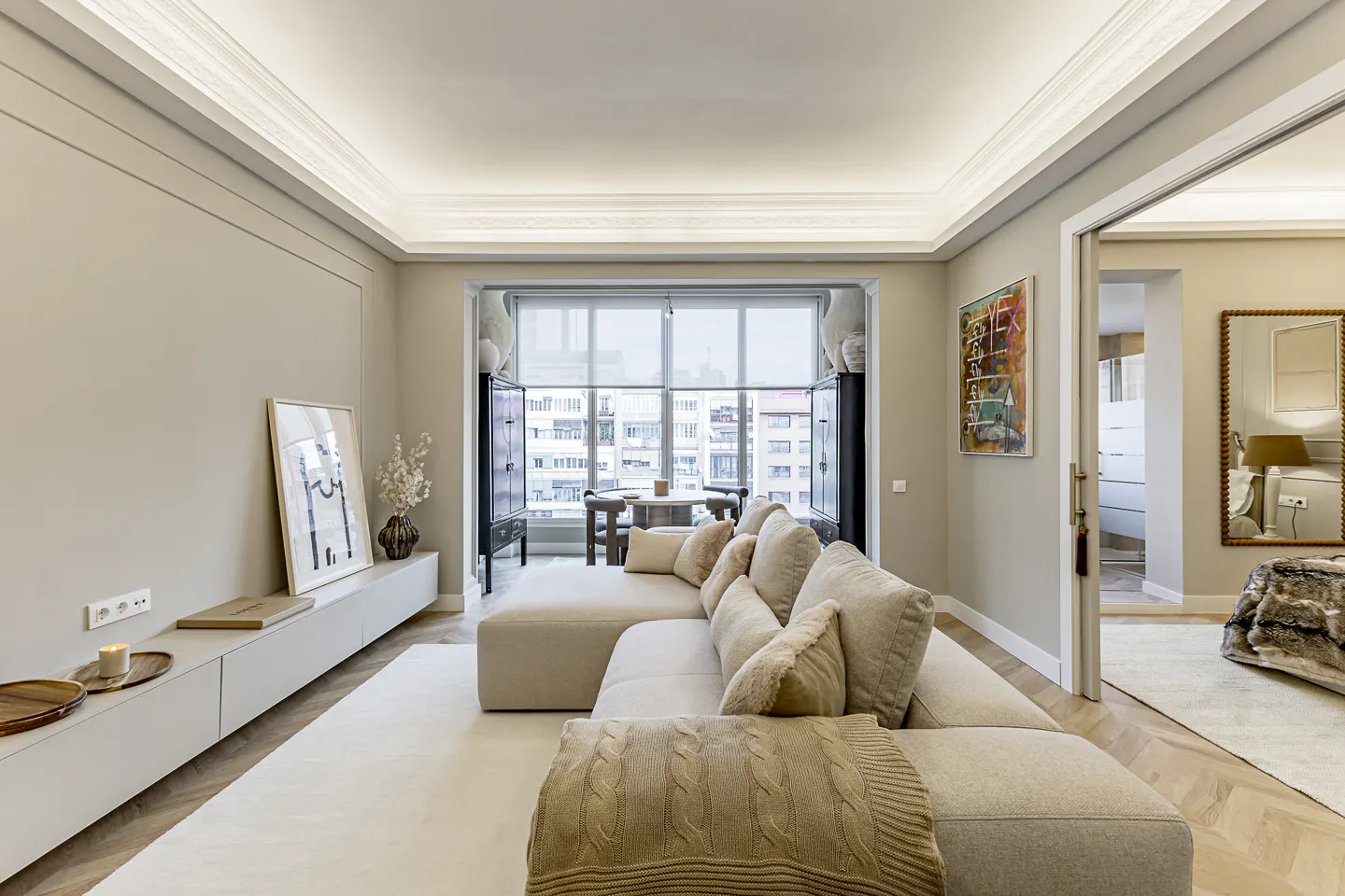 Living room with beige sectional sofa, throw blanket, and pillows. A black cabinet frames a window with city views. Artwork and decor add detail.