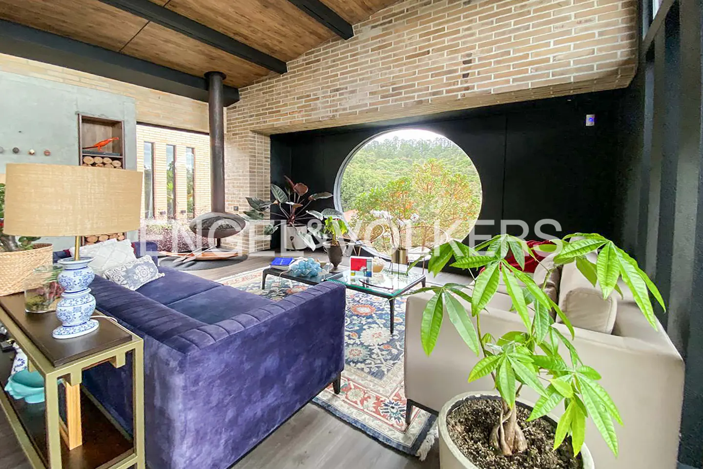 A modern living room with a blue sofa, plants, and a round window overlooking a green landscape.