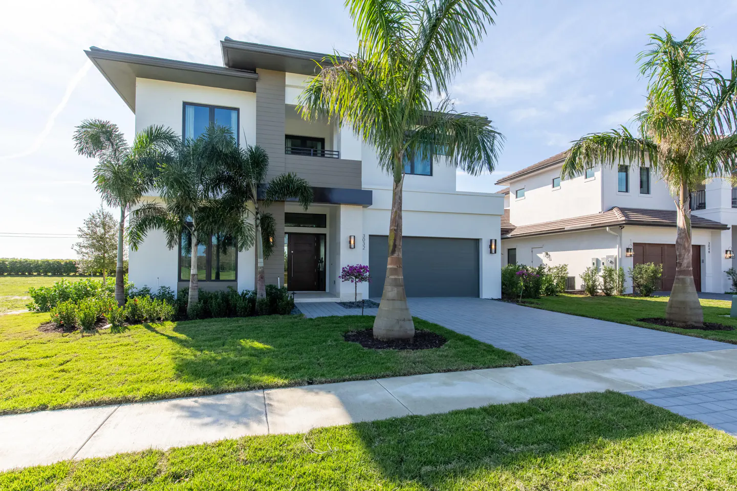 Modern two-story home with white and gray exterior, dark gray garage door, palm trees, and green lawn.