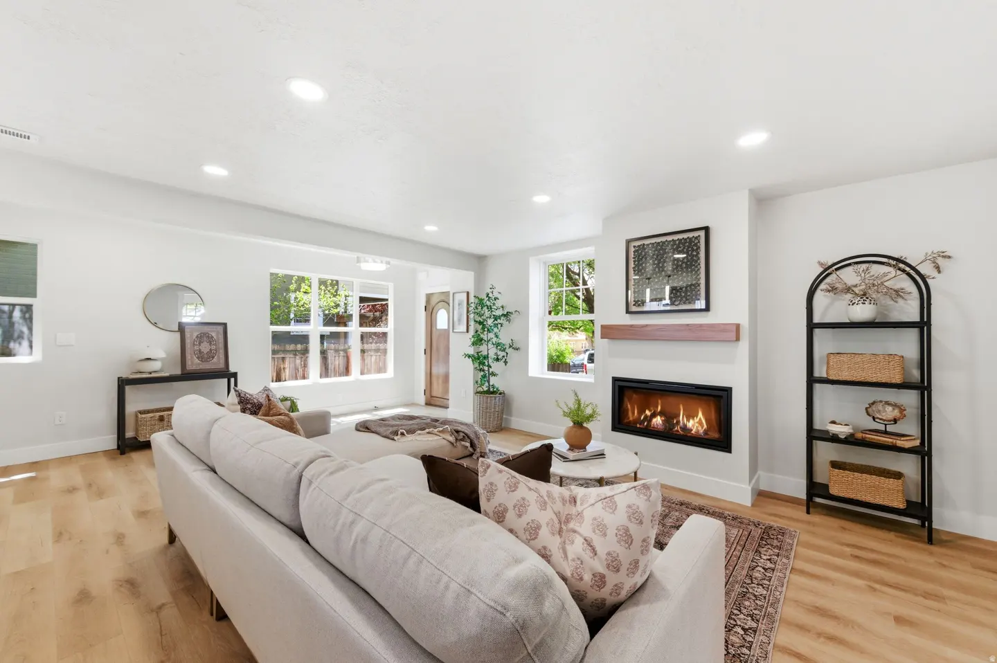 Bright, airy living room with a white sofa, fireplace, and wood floors. Windows offer a view of the outdoors.