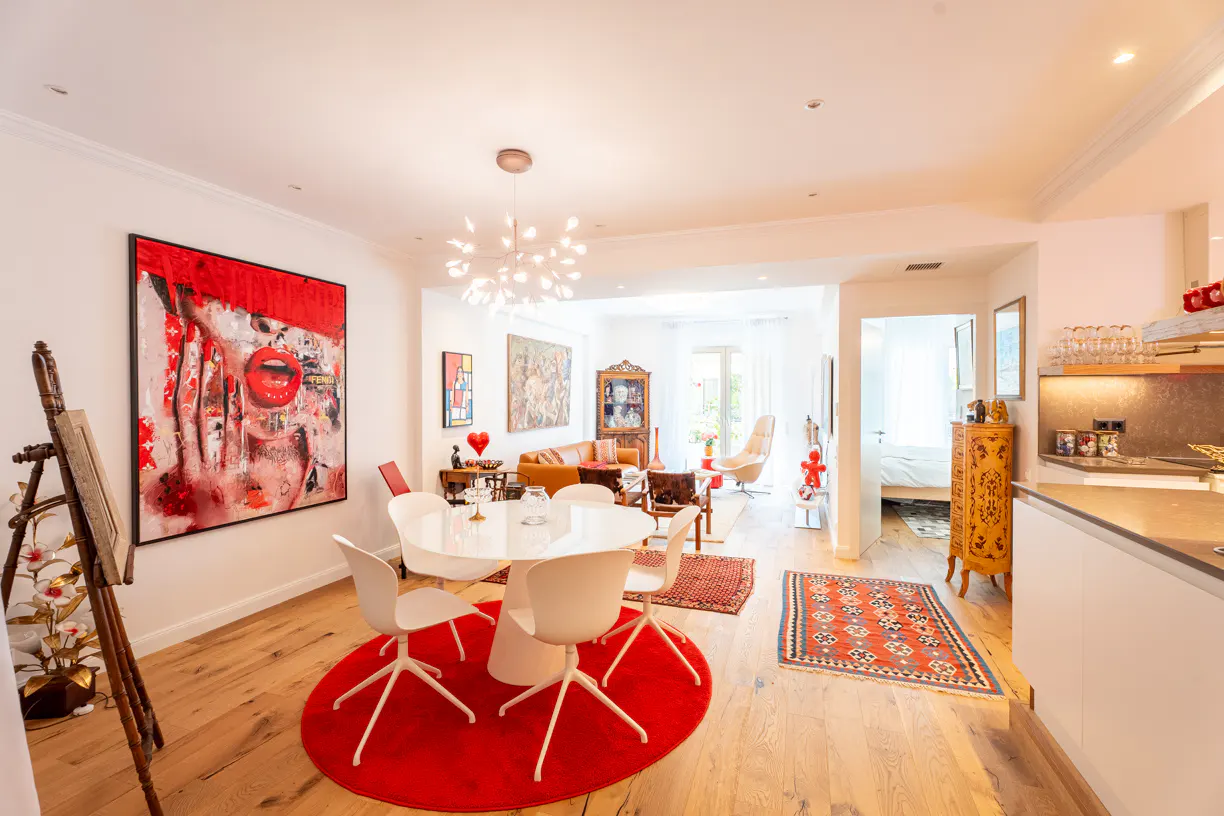 Bright, open-concept living space with white walls, wood floors, and colorful art. A round white table sits on a red rug.