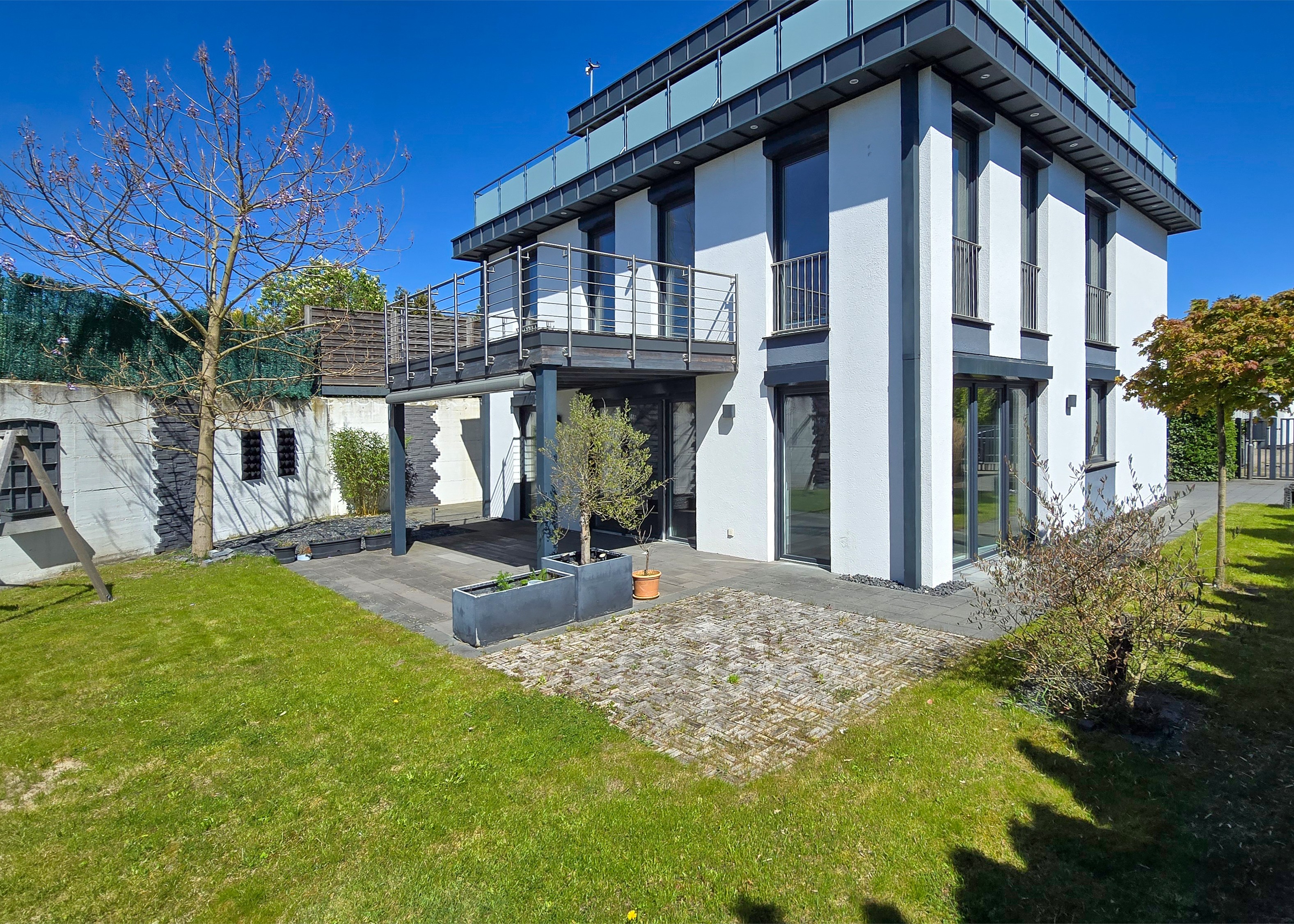 Modern white house with a balcony, dark trim, and large windows, surrounded by a green lawn and trees under a blue sky.