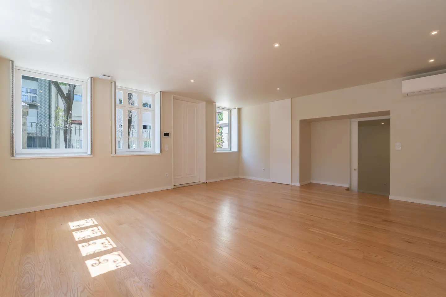 Bright, empty room with light wood floors, cream walls, and white trim. Three windows let in natural light.