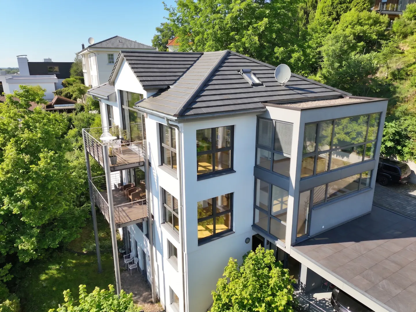 Modern white multi-story home with dark roof, large windows, stacked balconies, and lush green trees in a sunny setting.