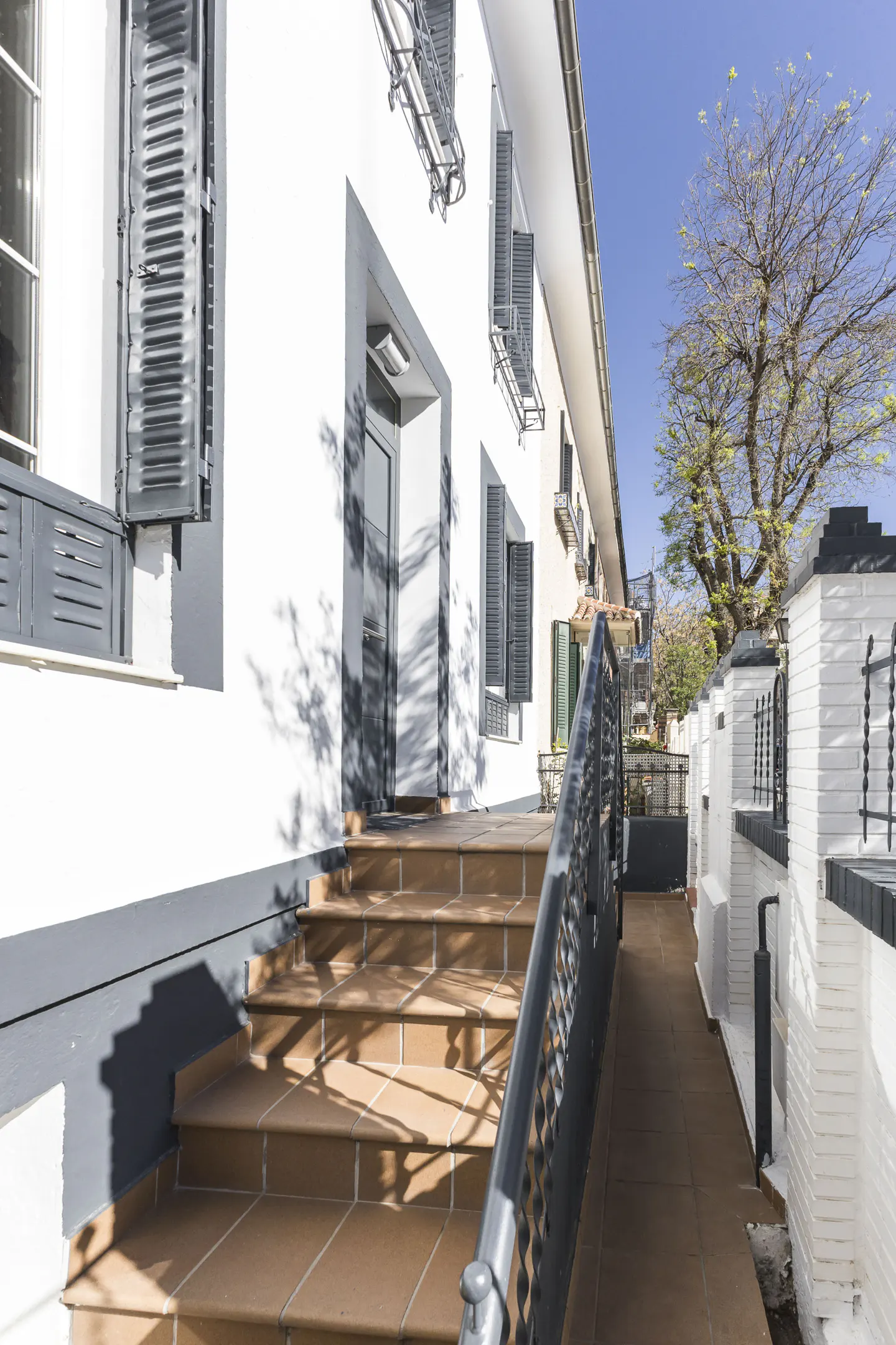 Exterior view of a white building with gray trim, brown tile steps, and a black metal railing. A tree is visible in the background.