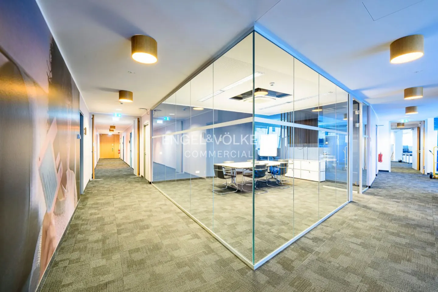 Office hallway with glass-walled Engel & Völkers Commercial conference room. Gray carpet, wood lights, and a mural on the left wall.