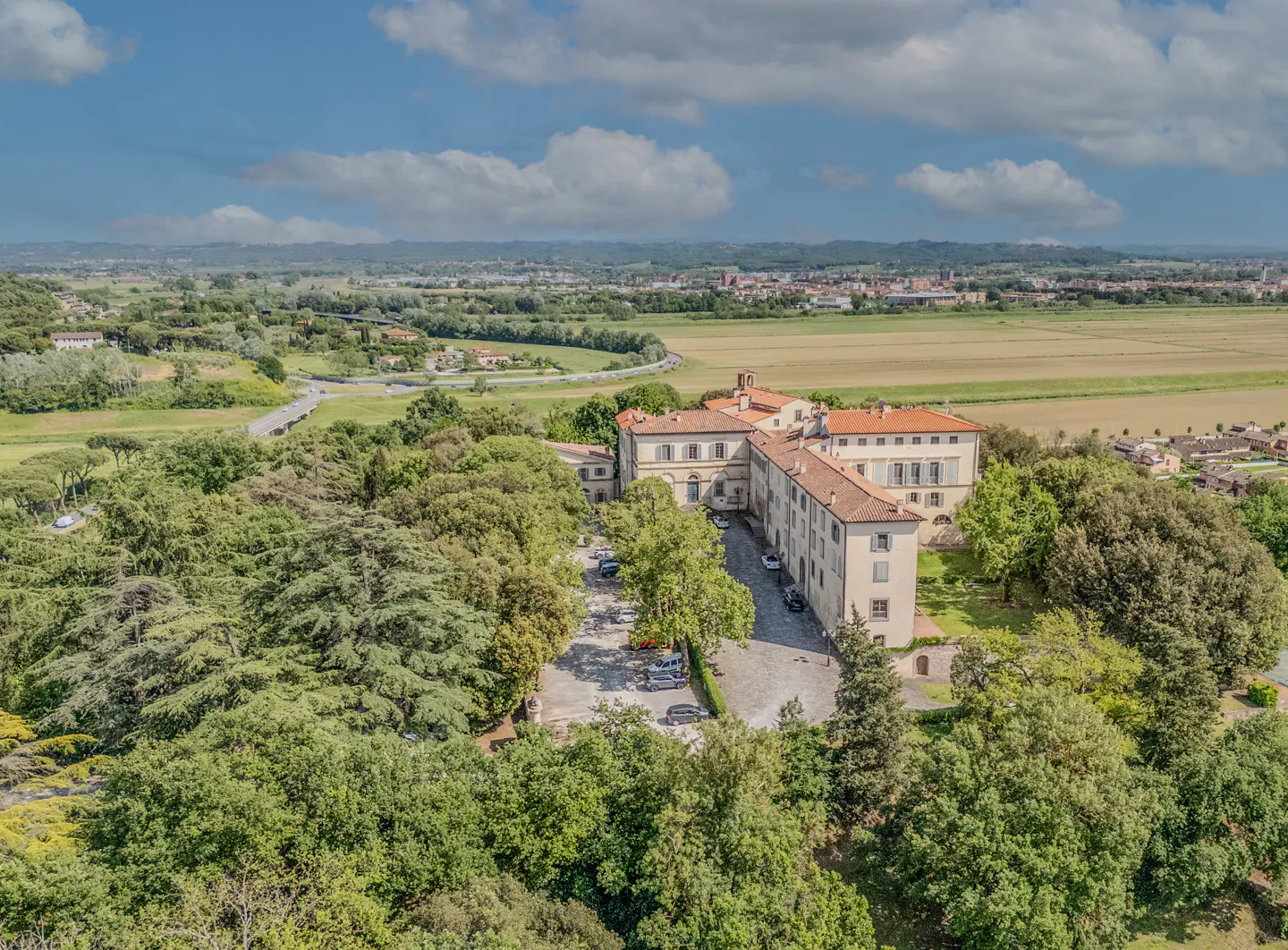 Aerial view of a large, light-colored building with a red tile roof, surrounded by green trees and fields under a blue sky with clouds.