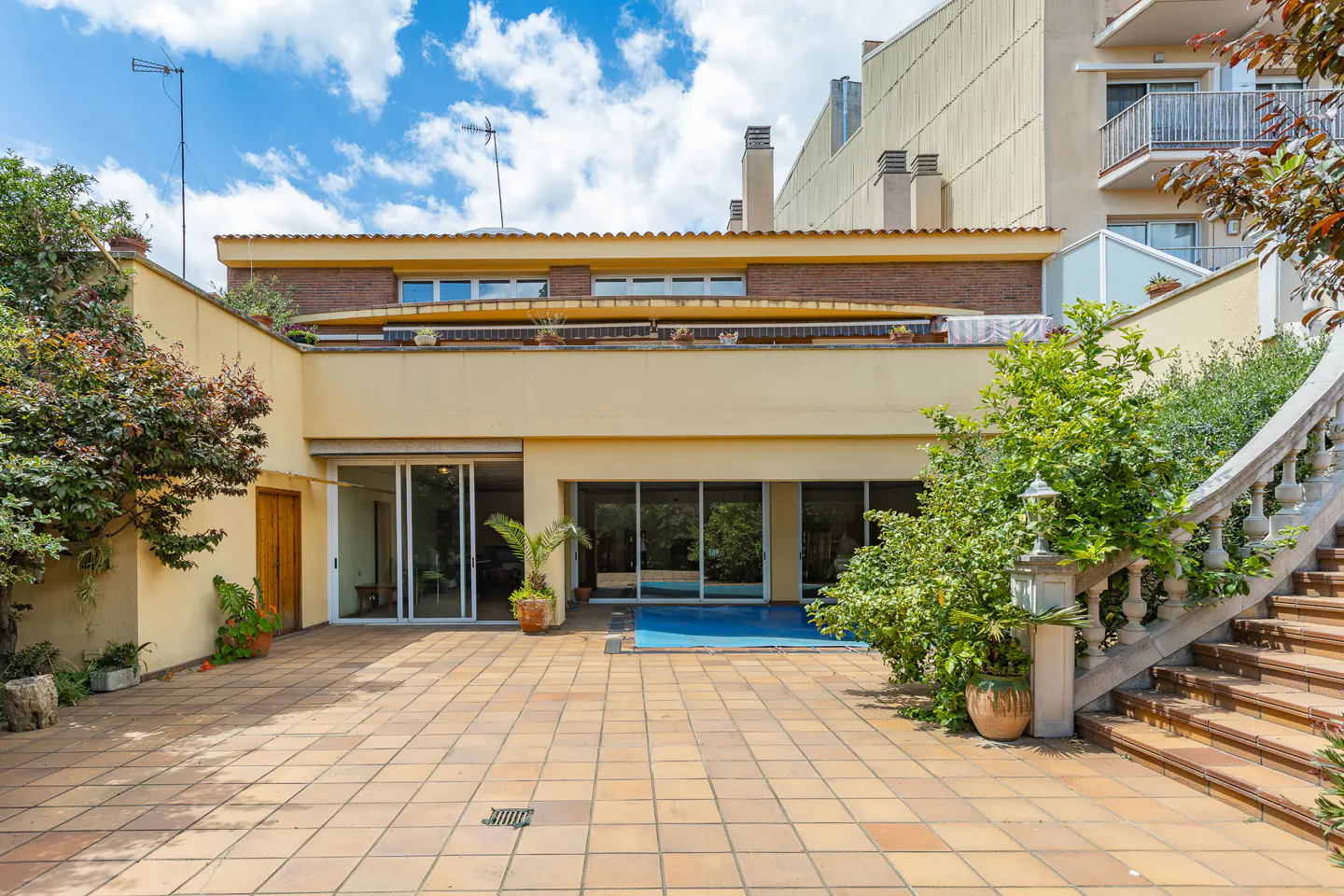 Backyard view of a two-story beige house with sliding glass doors, a tiled patio, and a staircase with a stone railing.