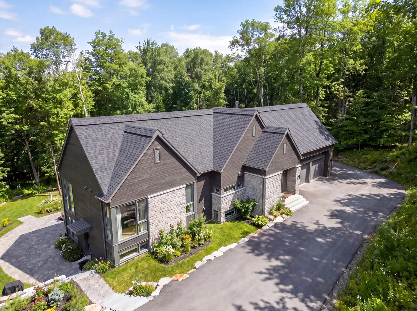 Aerial view of a modern, two-story home with a gray roof, stone accents, and a long driveway surrounded by lush green trees.