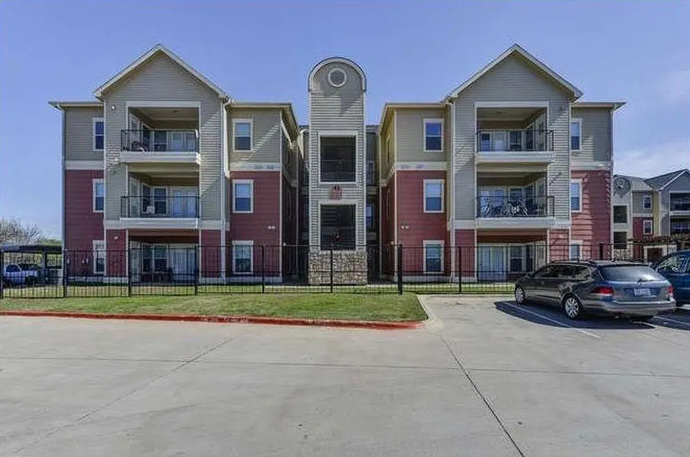 Three-story apartment building with red and beige siding, balconies, and a parking lot in front.
