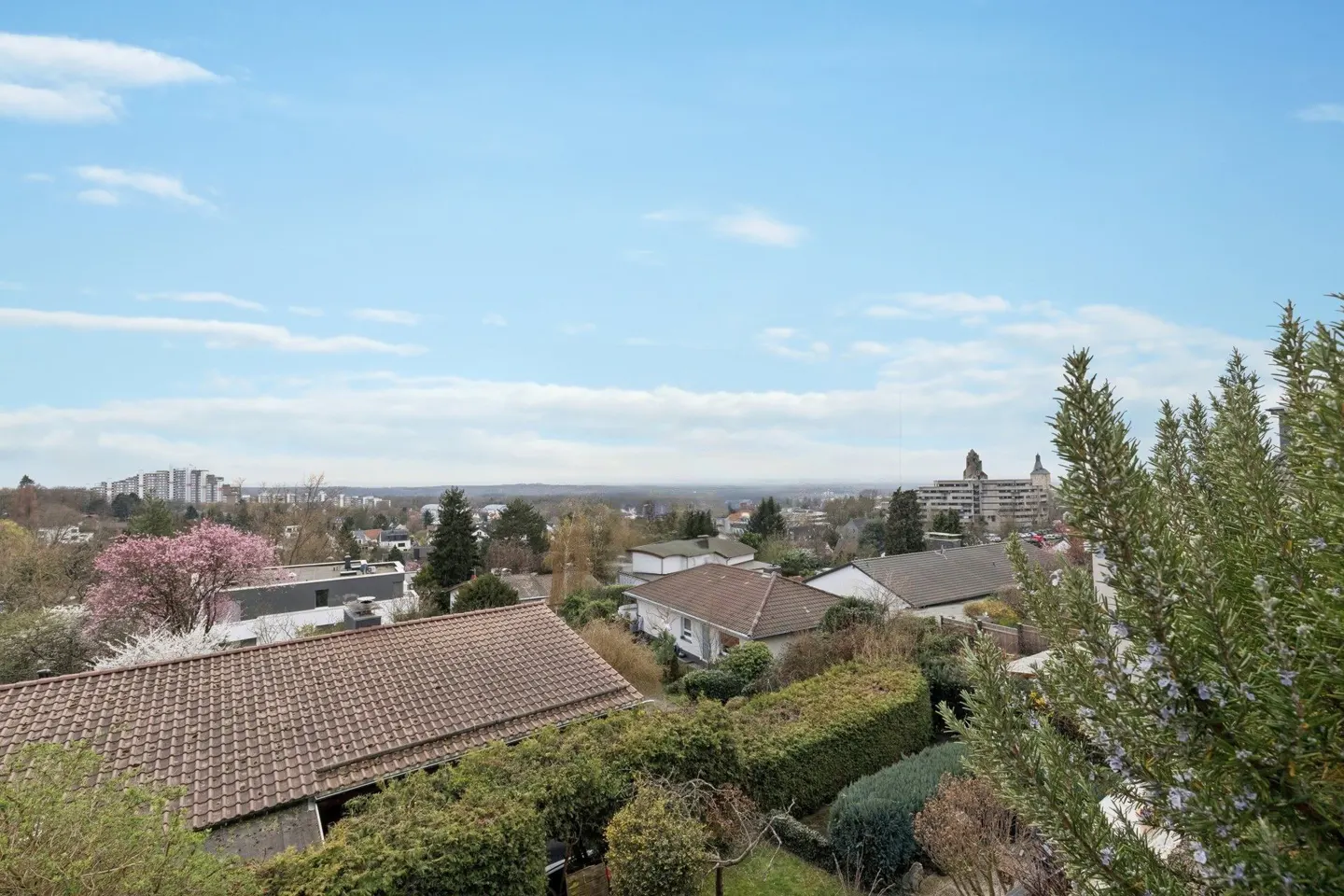 View of a residential area with houses, trees, and a blue sky with light clouds. In the distance, there are apartment buildings.