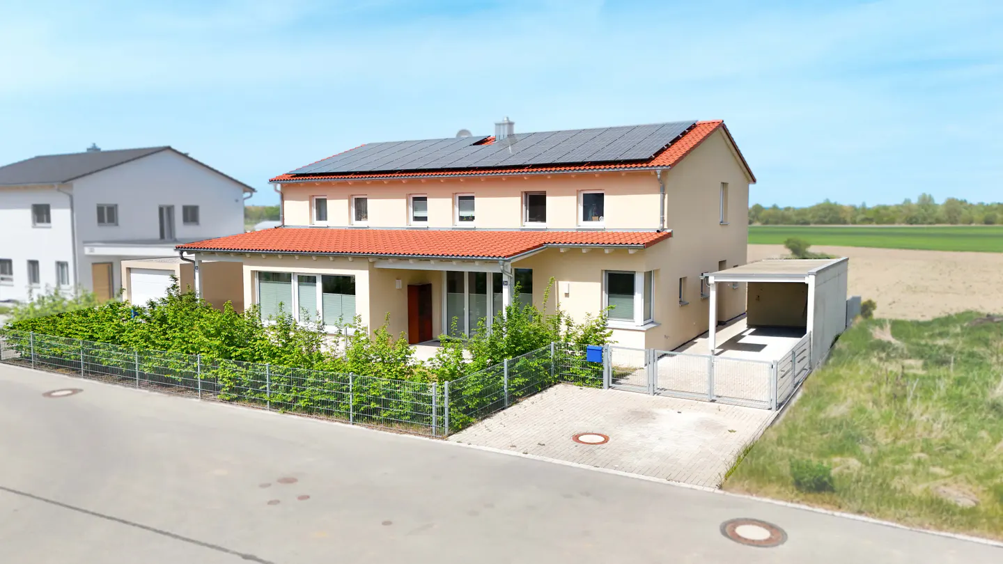 Two-story beige house with red tile roof and solar panels, green bushes, and a carport on a sunny day.