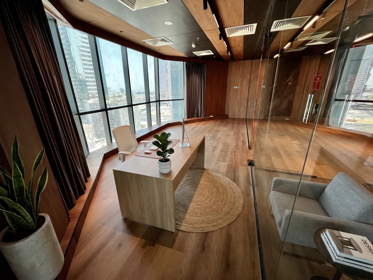 Office space with wood floors, a desk with a plant, and a view of the city through large windows. A gray sofa is visible through a glass wall.