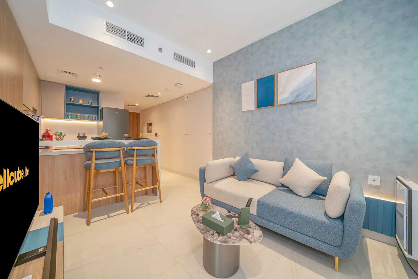 Modern apartment interior with blue accents. A sofa, bar stools, and a kitchen area are visible. Artwork adorns the blue wall.