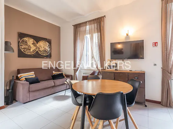 Modern living room with a brown sofa, round wooden dining table, black chairs, wall-mounted TV, and world map wall art.