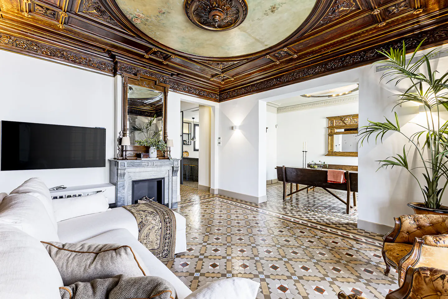 Living room with white sofa, marble fireplace, and ornate ceiling with a painted dome. Patterned tile floor and doorway to another room.