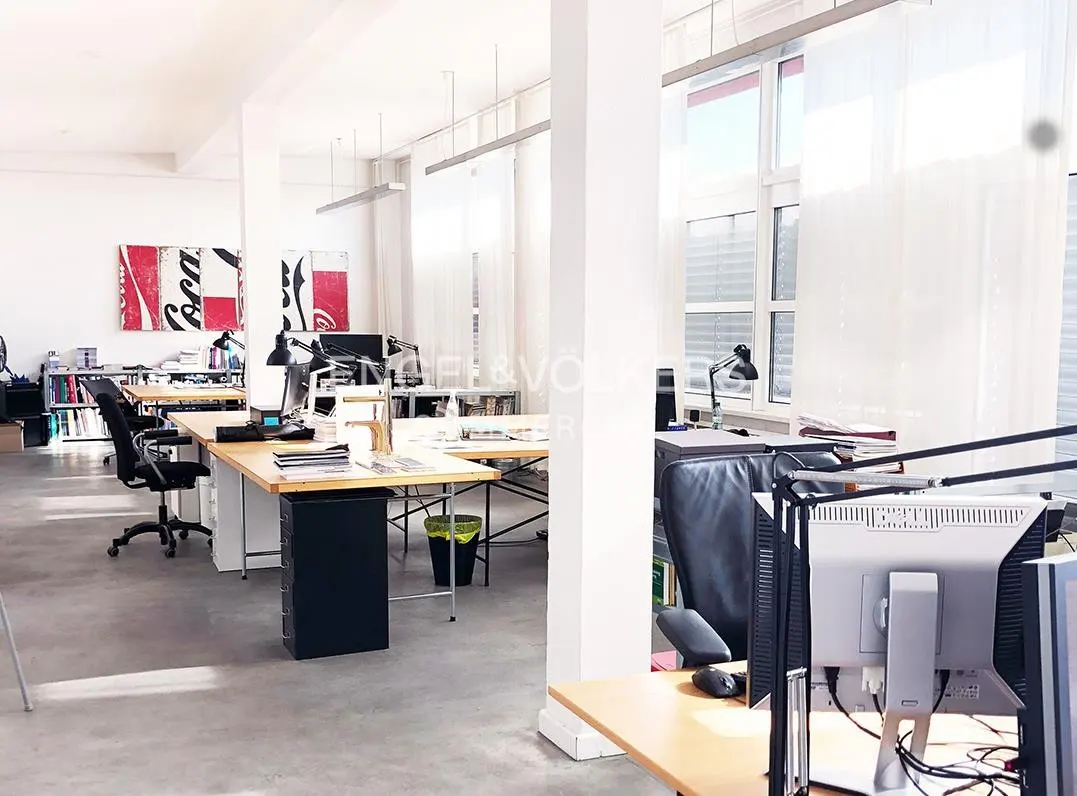 Bright, modern open-plan office with light wood desks, black chairs, large windows, and Coca-Cola wall art.