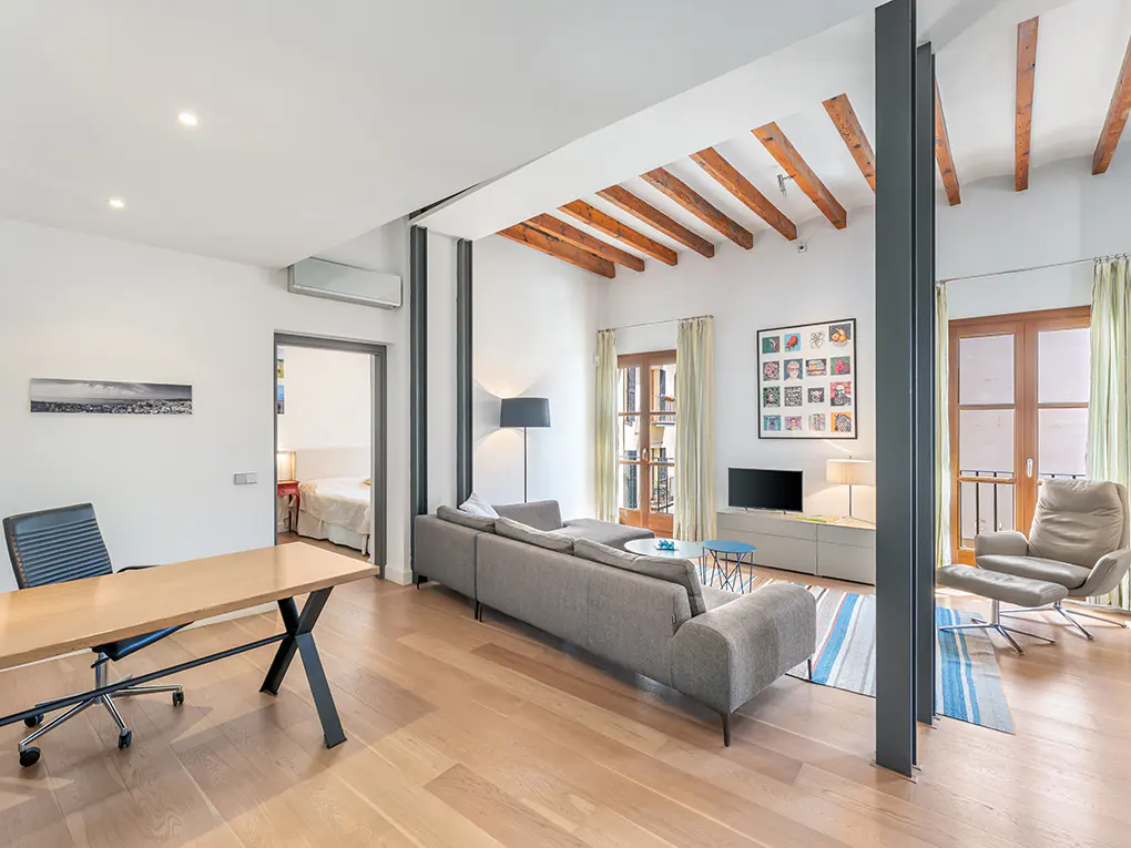 Bright, modern living room with wood floors, exposed beams, gray sofa, chair, and desk. Balcony visible through French doors.