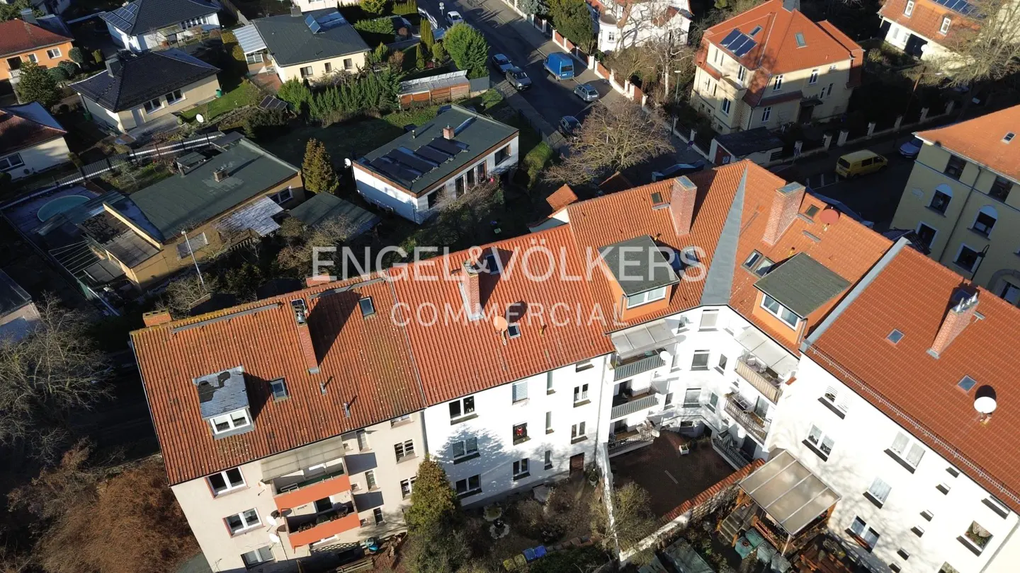 Aerial view of a white apartment building with a red tile roof, surrounded by trees and other houses. Engel & Völkers logo visible.