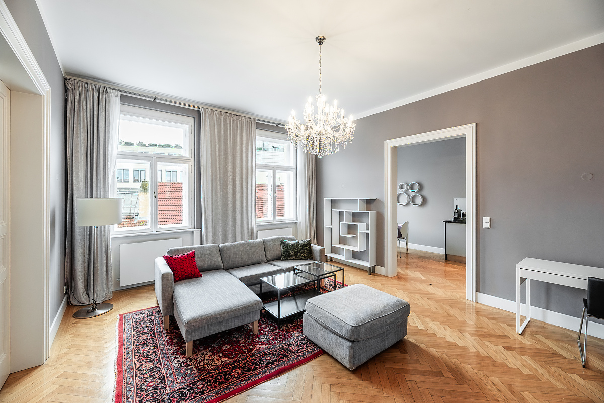 Modern living room with grey walls, hardwood floors, a grey sectional sofa, red accent pillow, and a crystal chandelier. [AI-generated text]