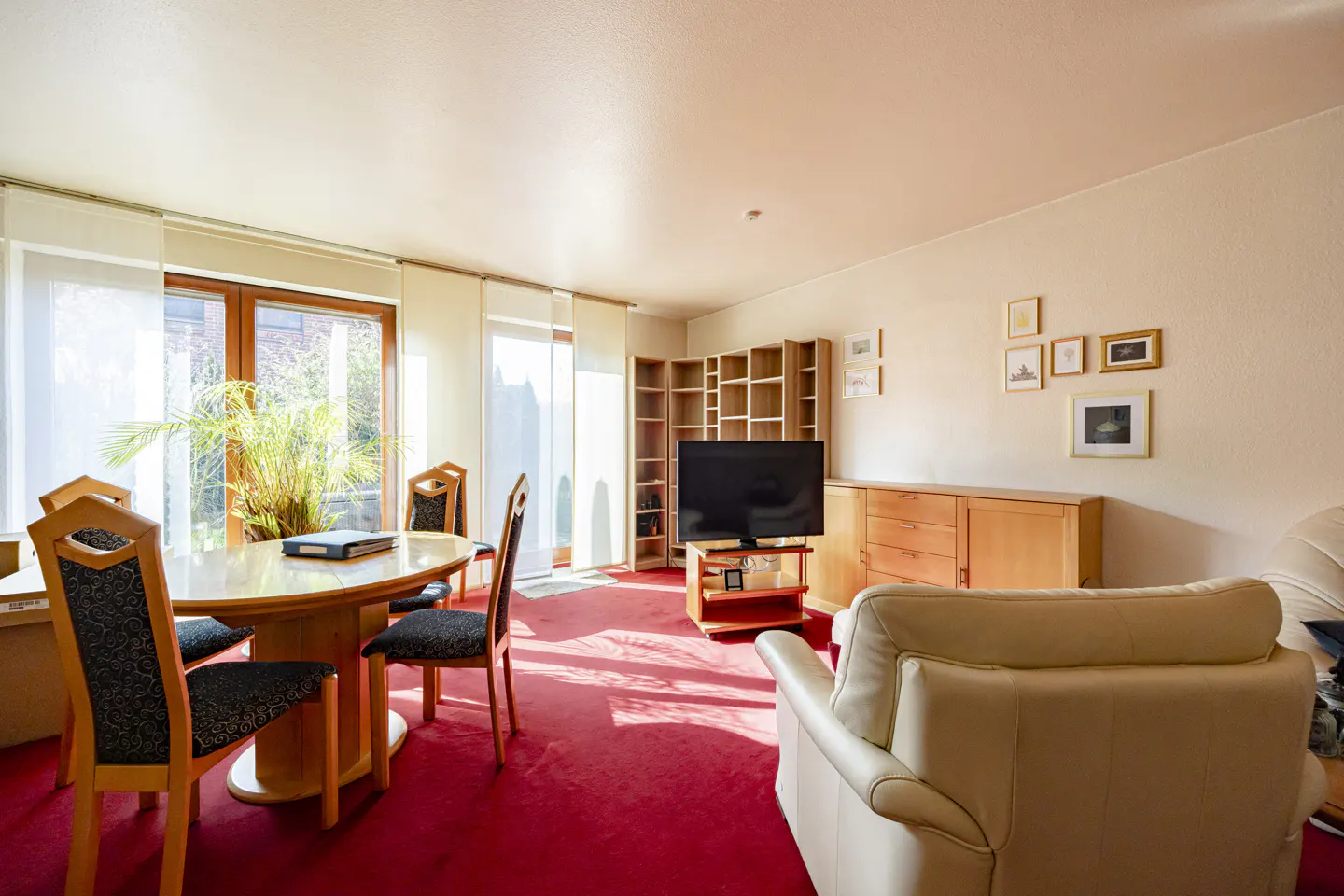Bright living room with red carpet, wooden furniture, and a cream-colored sofa. A dining table sits near a window with a view of greenery.