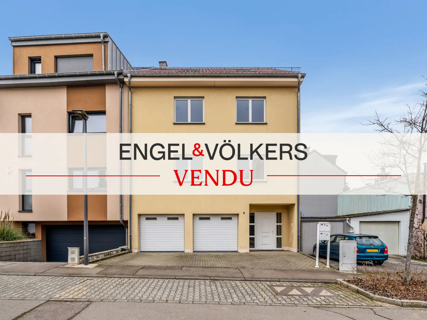 Exterior of a two-story tan house with two garage doors and an Engel & Völkers "Vendu" banner. A blue car is parked nearby.