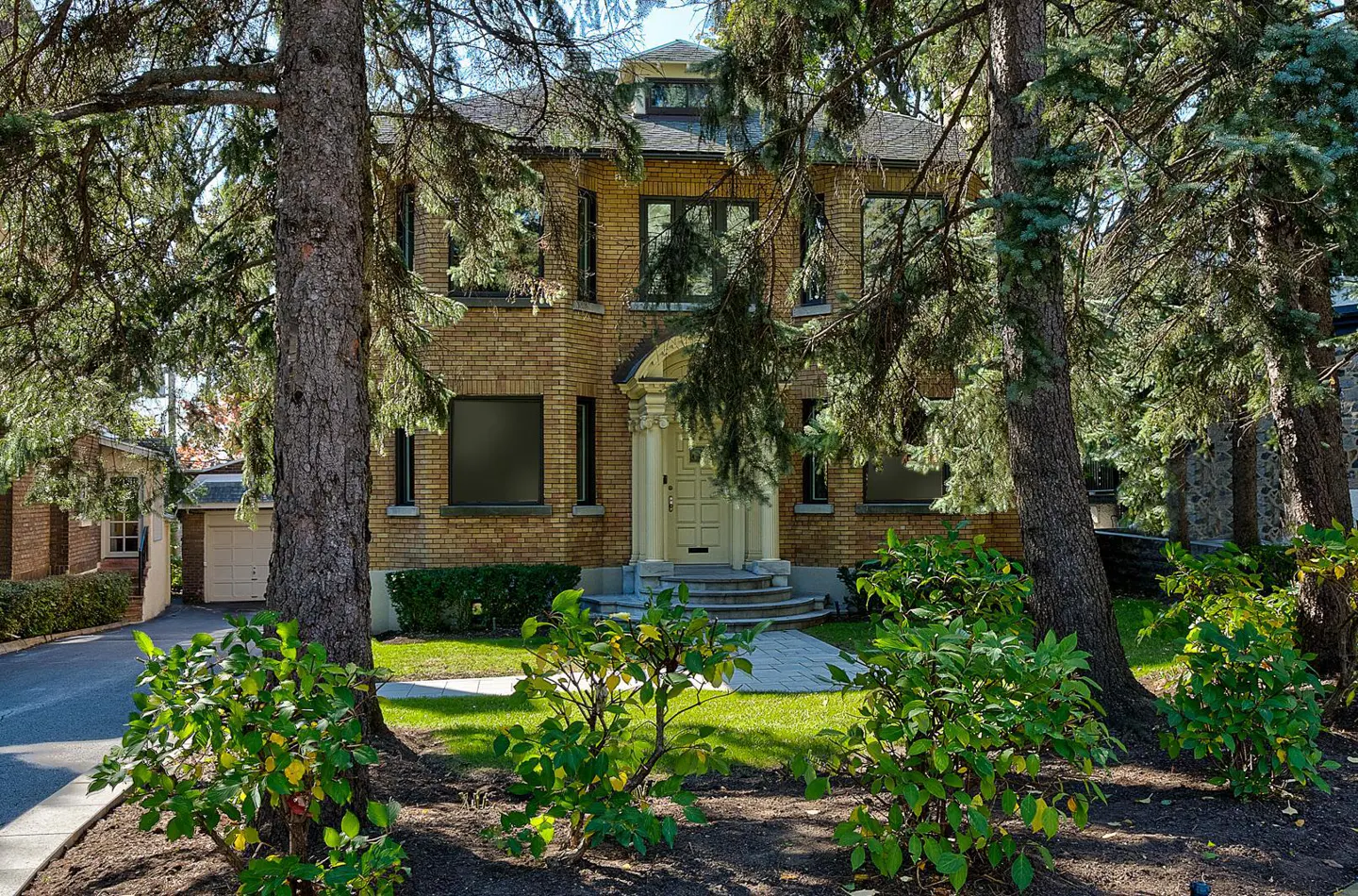 Two-story brick house with a light-colored front door and dark-framed windows, partially obscured by trees and green bushes.