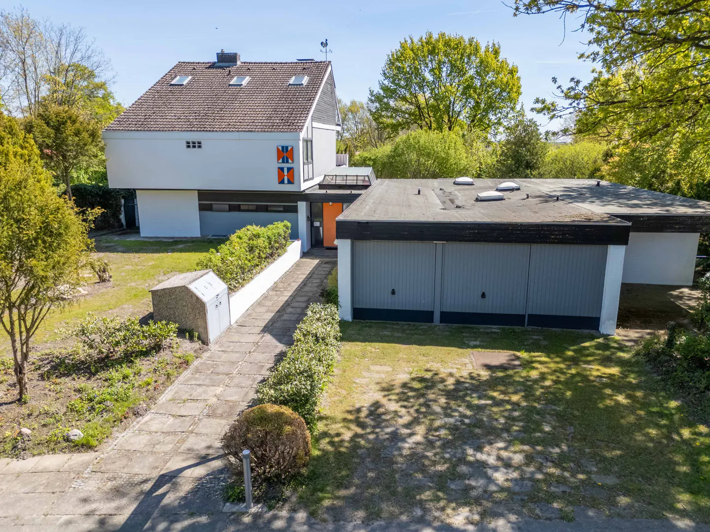 Modern white house with a brown roof, orange door, and gray garage doors, surrounded by green trees and a stone walkway.