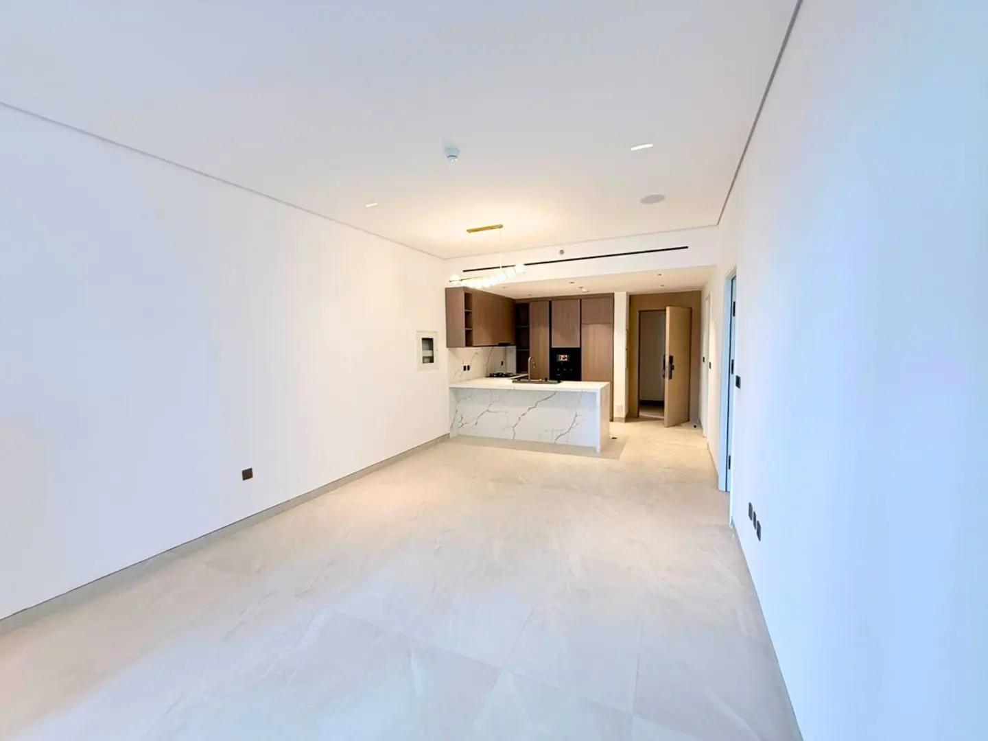 Bright, empty apartment with white walls and tile floors. A kitchen with wood cabinets and a marble island is in the background.