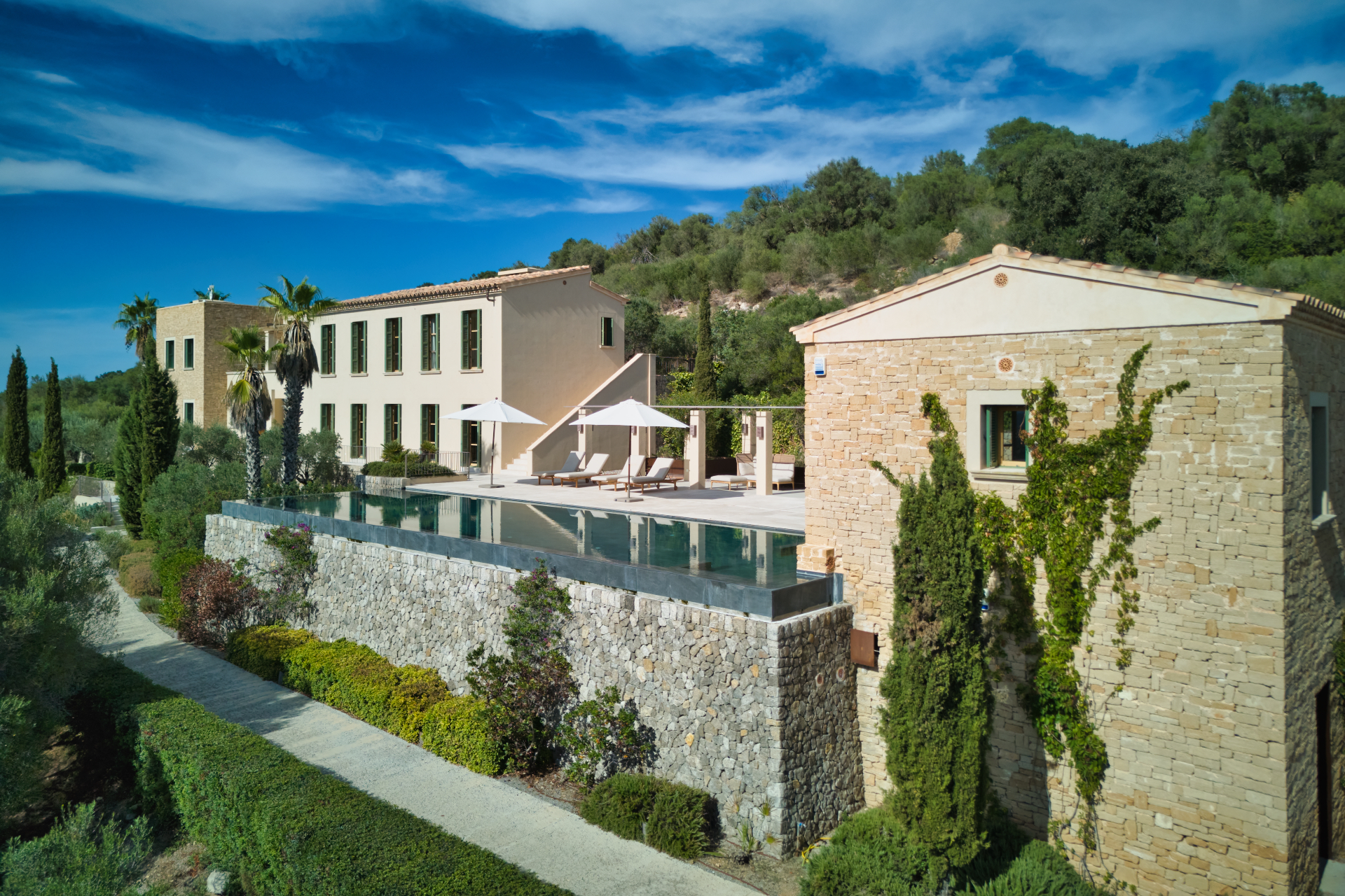 Luxury stone villa with an infinity pool, sun loungers, and umbrellas, set against a lush hillside under a blue sky. [AI-generated text]