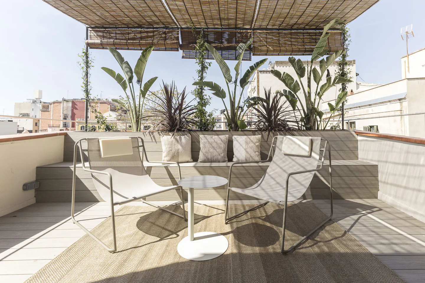 Rooftop patio with two chairs, a small table, and a built-in bench with pillows. Plants and a bamboo shade structure add greenery and shade.