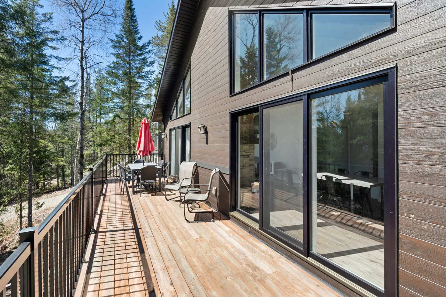Exterior view of a brown house with a wooden deck, black railings, and a red umbrella over a table and chairs.