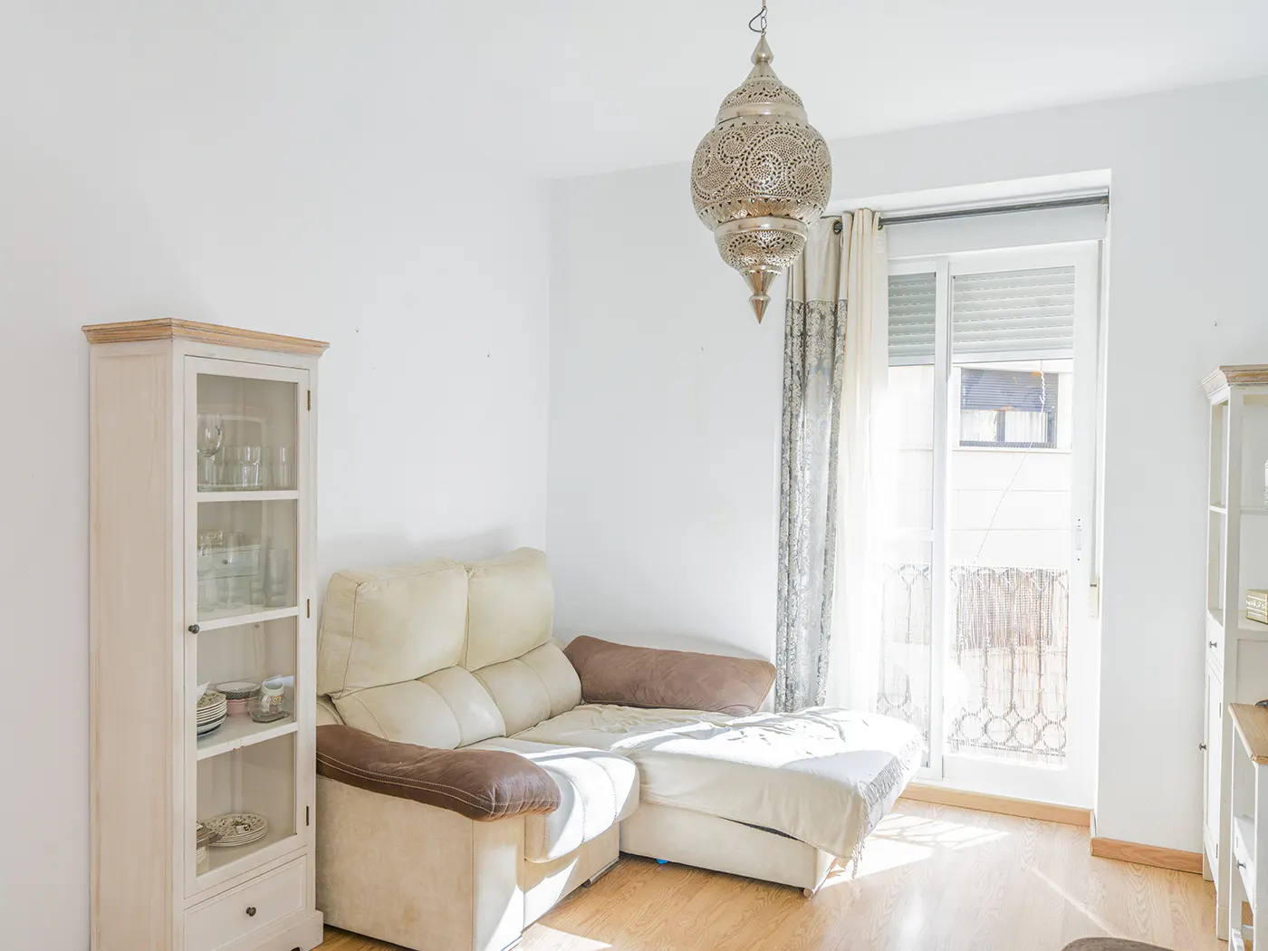 Bright living room with white walls, wood floors, and a beige sectional sofa. An ornate metal lamp hangs near a window with patterned curtains. A glass-front cabinet stands against the wall.