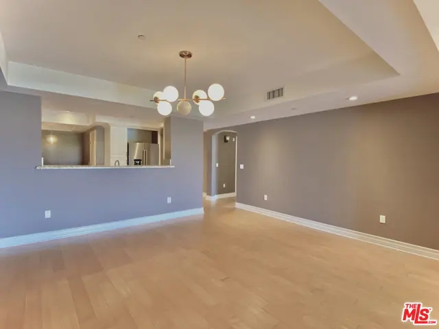A bright, empty living room with hardwood floors, gray walls, and a modern chandelier. A kitchen is visible through a cutout in the wall.