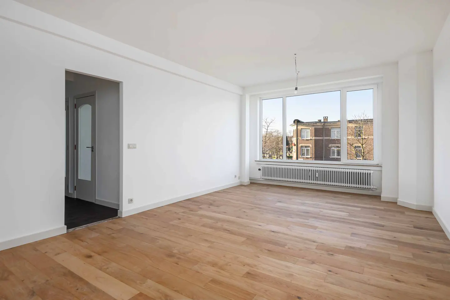 Bright, empty room with hardwood floors, white walls, and a large window showing a brick building. A doorway is visible on the left.