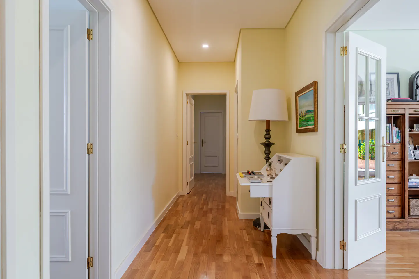 Hallway with light yellow walls, wood floors, and white trim. A white desk with a lamp sits against the wall. Open doors lead to other rooms.