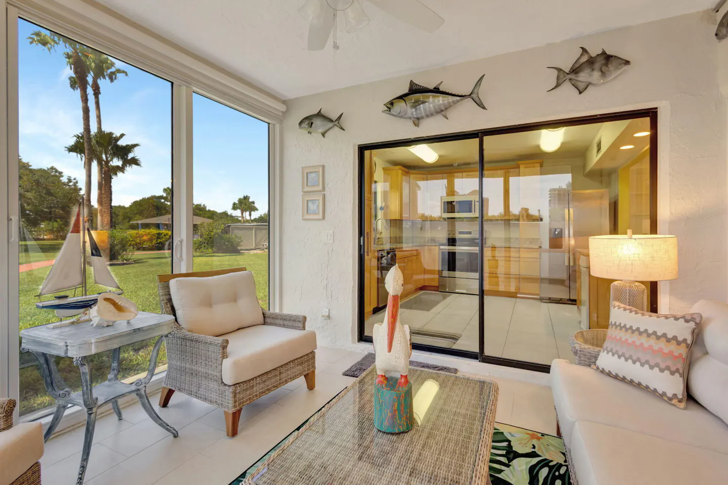 Sunroom with wicker furniture, fish decor, and a view of a green lawn with palm trees. Sliding glass doors lead to a kitchen.