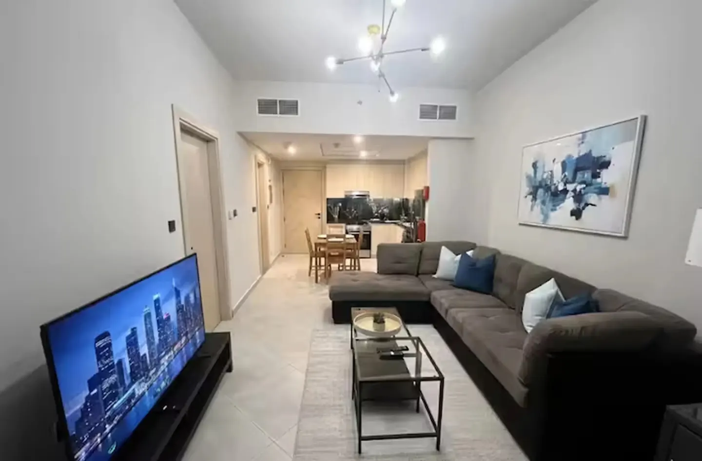 Living room with a gray sectional sofa, blue pillows, a glass coffee table, and a TV showing a city skyline. The kitchen is visible in the background.