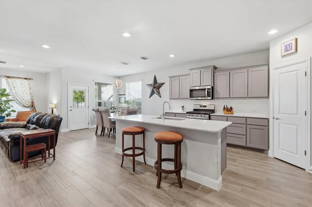 Open-concept living space with gray cabinets, white countertops, and wood-look tile flooring. Two stools at the kitchen island.